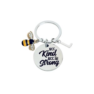 May include: A silver keychain with a round charm that reads "BEE KIND BEE STRONG." The keychain also features a small bee charm with black, yellow, and gold accents, and a small silver charm. The background is white.