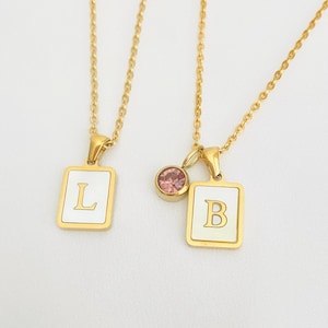May include: Two gold-colored necklaces with rectangular pendants. One pendant displays the letter "L", the other "B". Both pendants have a white background and gold trim. A small pink gemstone hangs from the chain of the "B" pendant.