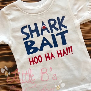 May include: White t-shirt with blue and red text that says "Shark Bait Hoo Ha Ha!!" and a small bee graphic. The text "Little B's Boy & Things" is written in pink below the graphic.