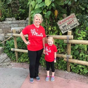 Puede incluir: Una madre y su hija llevan camisetas rojas con gr&aacute;ficos de Minnie Mouse. La camiseta de la madre dice "Me Minnie" y la camiseta de la hija dice "Minnie Me". Est&aacute;n de pie frente a una valla de madera con un cartel que dice "&iexcl;Advertencia! No tire de la cuerda".