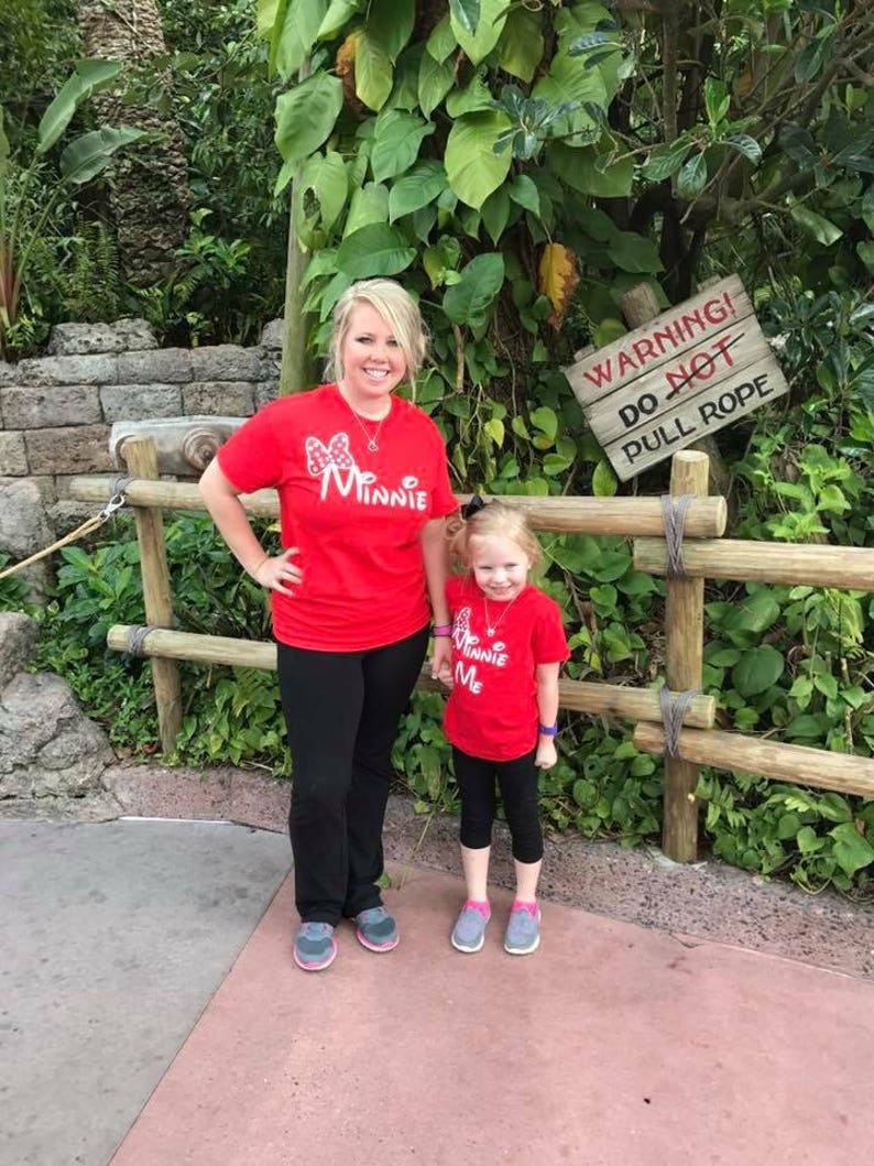 Puede incluir: Una madre y su hija llevan camisetas rojas con gr&aacute;ficos de Minnie Mouse. La camiseta de la madre dice "Me Minnie" y la camiseta de la hija dice "Minnie Me". Est&aacute;n de pie frente a una valla de madera con un cartel que dice "&iexcl;Advertencia! No tire de la cuerda".