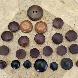 May include: A collection of 20 wooden buttons in various shades of brown, black, and beige. Some buttons have a swirl pattern, while others are plain. The buttons are arranged in a triangular shape on a light brown surface.