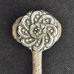 May include: A silver-toned metal button with a floral design. The button is round and has a raised center with a swirl pattern. The button is attached to a metal stem.
