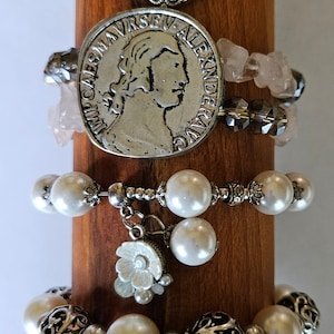 May include: A wooden display stand with five silver bracelets. The bracelets feature various charms, including a heart, a coin, a flower, and a cross. The charms are all silver and have intricate designs.