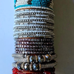 May include: A stack of colorful beaded bracelets, including turquoise, silver, purple, gray, red, and cream. The bracelets are stacked on a wooden dowel. The top bracelet has a turquoise and white beaded pattern with a silver accent. The label on the top bracelet reads "Style x Muse Bracelet".