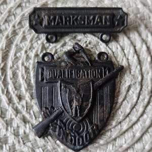 May include: Two antique metal badges. The top badge reads "MARKSMAN" with star accents. The bottom badge is shield-shaped, with the word "QUALIFICATION" and a rifle design. Both badges are dark brown with signs of wear.