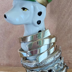 May include: A white ceramic dalmatian dog figurine wearing a green and gold hat, holding a stack of silver bracelets.