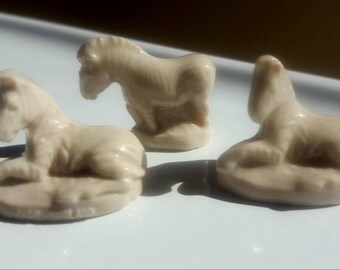 May include: Three ceramic figurines of zebras, each in a different pose. The zebras are a light beige color and are sitting on a white surface.