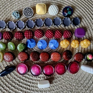 May include: A collection of various buttons in different shapes, sizes, and colors. The buttons include round, square, and textured designs in shades of blue, red, brown, yellow, and green. Some buttons have a floral design.