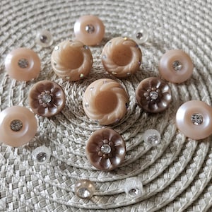 May include: A collection of decorative buttons in various shades of brown and beige, some with a swirl design, others with a floral pattern. Several buttons have a central rhinestone. Clear, small rhinestones are scattered around the buttons.