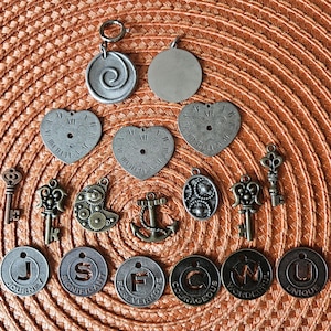 May include: A collection of antique-style charms and pendants in various shapes and designs. Includes heart-shaped clock faces, keys, and circular charms with words like "Journey" and "Unique". The color palette is primarily metallic silver and bronze.