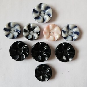 May include: A collection of decorative buttons in various colors and designs. The buttons include black, white with blue swirls, and a light pink button. Each button has two holes for sewing.