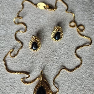 May include: A gold-toned jewelry set featuring a necklace with a teardrop pendant, and matching teardrop earrings. Each piece has a black stone center. The necklace chain and earring frames have a detailed design.