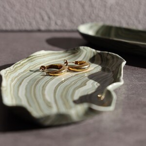 May include: A small, irregular, oval-shaped tray featuring a marbled pattern of sage green, cream, and dark brown swirls. Resting on the surface of the tray are two polished gold-toned hoop earrings with a smooth, reflective metallic finish.
