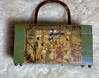May include: A rectangular olive green wooden box with a brown arched handle and a metal latch. The front panel features a detailed illustration of a 19th-century shop interior with figures, a coffee grinder, and a cat. The box stands on four small feet.