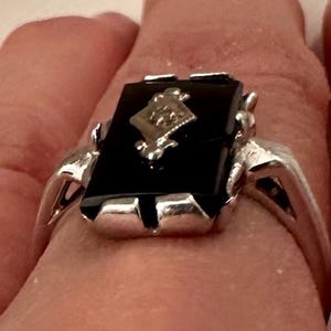May include: A silver ring with a rectangular black onyx stone. The ring features a diamond-shaped design in the center of the onyx and a silver band. The ring is a classic piece of jewelry.