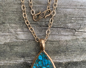 May include: A gold-coloured necklace featuring a teardrop-shaped pendant. The pendant is filled with a vibrant turquoise stone. The necklace chain and clasp are also gold-coloured, creating a cohesive design. The necklace is displayed on a wooden surface.
