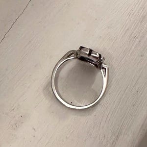 May include: A silver ring with a square-shaped, dark-colored gemstone. The ring has a simple band and a decorative setting around the stone. The ring is shown on a white surface.