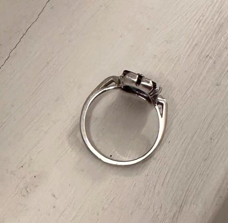 May include: A silver ring with a square-shaped, dark-colored gemstone. The ring has a simple band and a decorative setting around the stone. The ring is shown on a white surface.