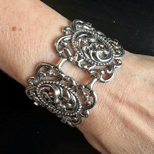 May include: Silver-toned filigree cuff bracelet. The bracelet features two ornate, oval-shaped panels connected by short bars. Each panel has a detailed, swirling design with beaded edges. The bracelet is worn on a wrist.