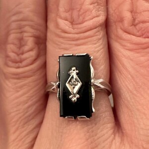 May include: A rectangular black onyx ring with a silver band. The ring features a diamond-shaped design with a small diamond in the center. The ring is set in a silver setting with decorative flourishes.