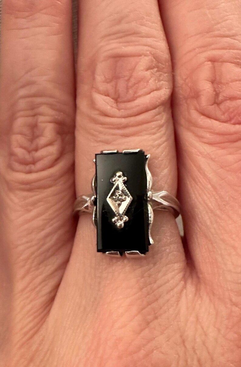 May include: A rectangular black onyx ring with a silver band. The ring features a diamond-shaped design with a small diamond in the center. The ring is set in a silver setting with decorative flourishes.