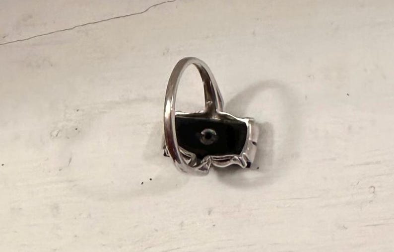 May include: A silver ring with a rectangular black stone and a small, round, clear stone in the center. The ring has a decorative, floral-like design around the black stone.