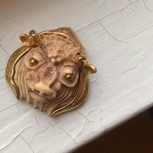 May include: Vintage brooch featuring a detailed lion's head design. The brooch has a cream-colored face with a textured mane and a gold-tone frame. Two small gold accents are on the top of the head. A unique piece of jewelry.