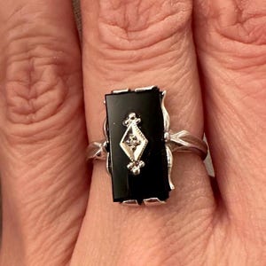 May include: A silver ring featuring a rectangular black onyx stone with a diamond-shaped accent. The ring has a decorative, ornate design on the sides and is worn on a finger.