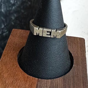May include: A silver-toned ring with the letters "MEM" prominently displayed. The ring is set against a black background and a wooden stand. The ring's design is simple and bold, with a focus on the letters.