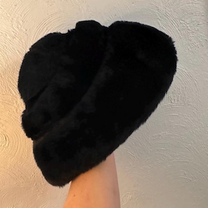 May include: A black faux fur hat with ear flaps. The hat is made of a soft, plush material and has a rounded crown. The ear flaps extend down to cover the ears. The hat is a solid black color.