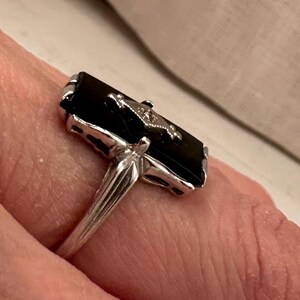 May include: A silver ring featuring a rectangular black onyx stone with a small, decorative accent. The ring's band is a simple, polished silver, and the setting around the onyx has intricate detailing.