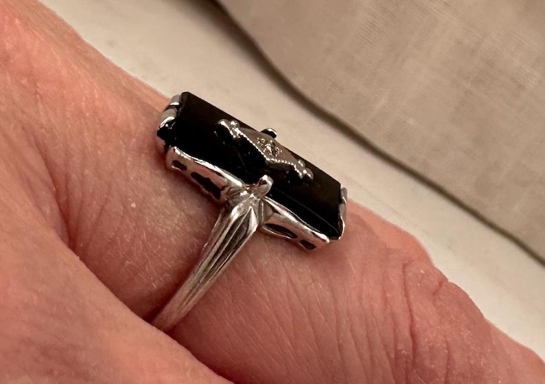 May include: A silver ring featuring a rectangular black onyx stone with a small, decorative accent. The ring's band is a simple, polished silver, and the setting around the onyx has intricate detailing.