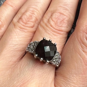 May include: A silver ring featuring a large, faceted, dark gemstone. The band is adorned with smaller, sparkling stones. The ring is displayed on a hand, showcasing its design and details. A piece of jewelry.