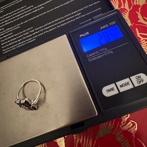 May include: A silver ring with a dark gemstone is placed on a digital scale. The scale displays a weight of 2.8 grams. The scale is black with white text and has buttons for TARE, MODE, ON, and OFF.