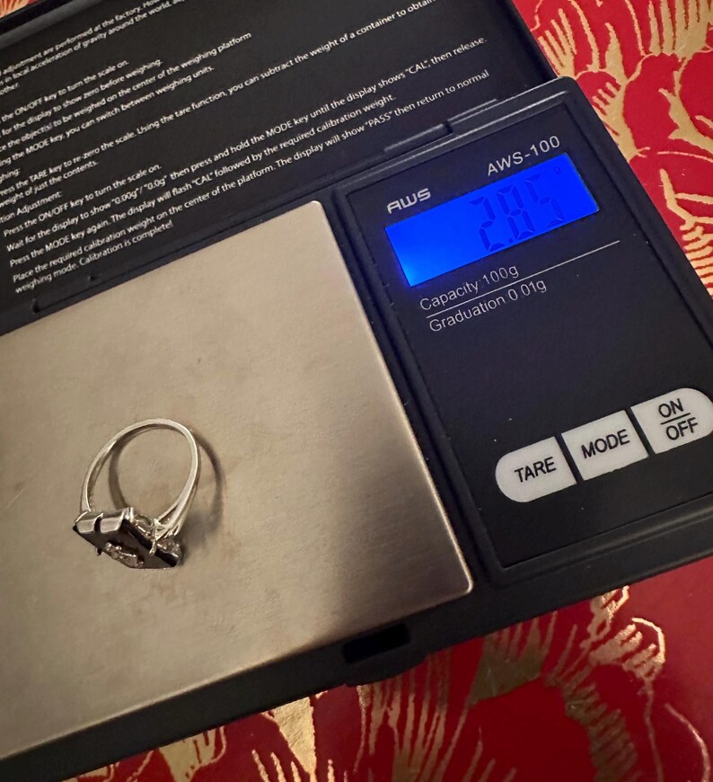May include: A silver ring with a dark gemstone is placed on a digital scale. The scale displays a weight of 2.8 grams. The scale is black with white text and has buttons for TARE, MODE, ON, and OFF.