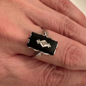 May include: A silver ring featuring a rectangular black onyx stone with a diamond-shaped, diamond-studded center. The ring has a decorative, scalloped edge and is set on a silver band.