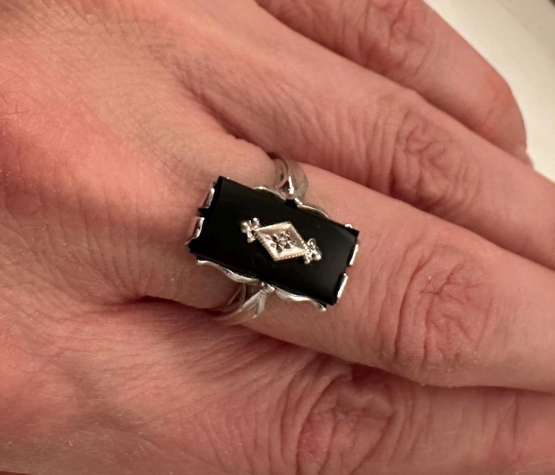 May include: A silver ring featuring a rectangular black onyx stone with a diamond-shaped, diamond-studded center. The ring has a decorative, scalloped edge and is set on a silver band.