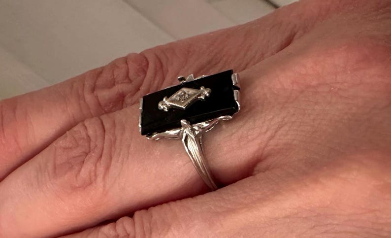 May include: A rectangular black onyx ring with a diamond-shaped accent in the center. The ring has a silver-colored band and decorative detailing. The ring is set on a finger.