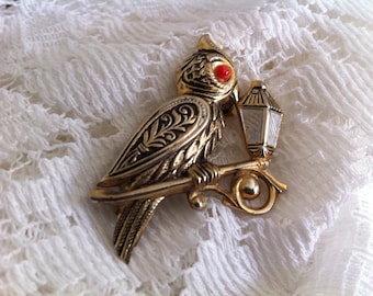 May include: A vintage gold-coloured owl brooch with a red eye and a small lantern. The owl is perched on a branch, and the brooch has intricate detailing. The background is a white, textured fabric.