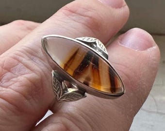 May include: Oval-shaped silver ring with a large, polished stone. The stone features brown and cream-colored bands. The ring has leaf-like details on the sides and top. A unique piece of jewelry.