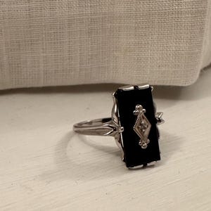 May include: A silver ring with a rectangular black onyx stone. The ring features a diamond-shaped design with a small diamond in the center. The ring is set on a white surface.