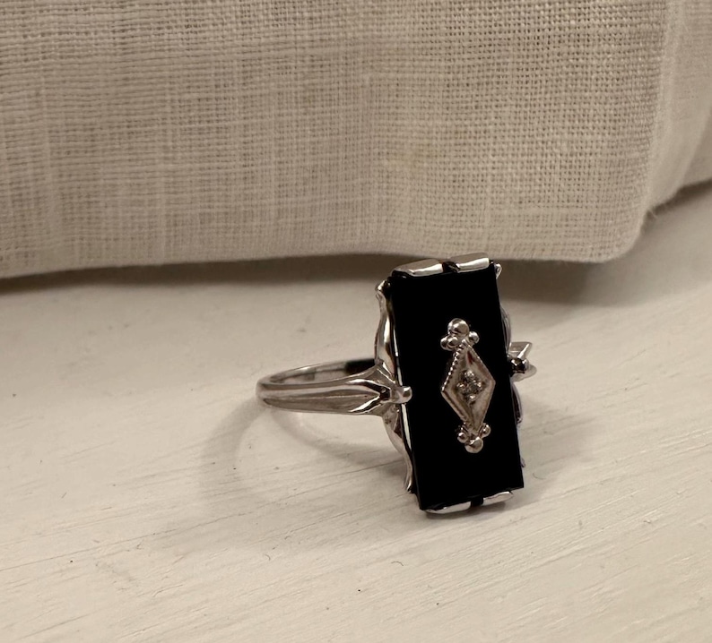 May include: A silver ring with a rectangular black onyx stone. The ring features a diamond-shaped design with a small diamond in the center. The ring is set on a white surface.