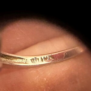 May include: Close-up of a gold ring with visible markings. The ring's band is slightly curved, and the markings appear to be stamps or engravings. The background is a blurred, warm-toned surface, likely skin.