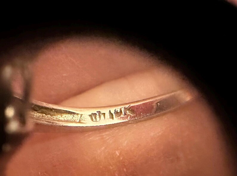May include: Close-up of a gold ring with visible markings. The ring's band is slightly curved, and the markings appear to be stamps or engravings. The background is a blurred, warm-toned surface, likely skin.