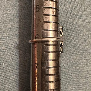 May include: A silver ring with two dark stones is measured on a ring sizer. The ring is placed on the sizer between sizes 6 and 7. The ring has a simple band and a decorative setting for the stones.