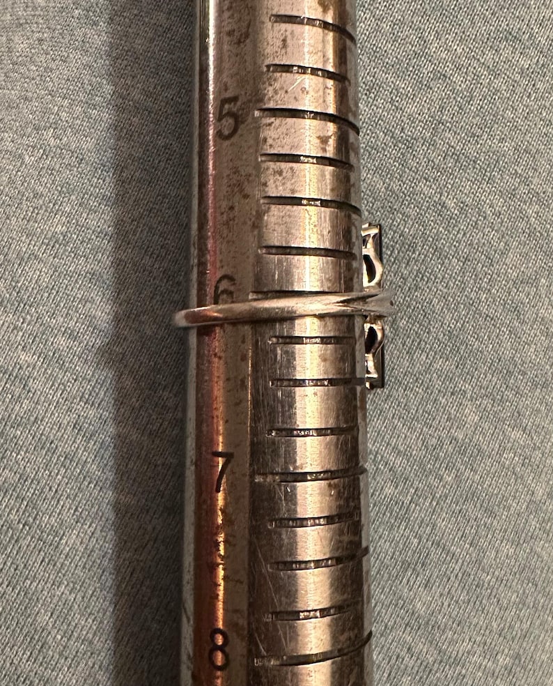 May include: A silver ring with two dark stones is measured on a ring sizer. The ring is placed on the sizer between sizes 6 and 7. The ring has a simple band and a decorative setting for the stones.