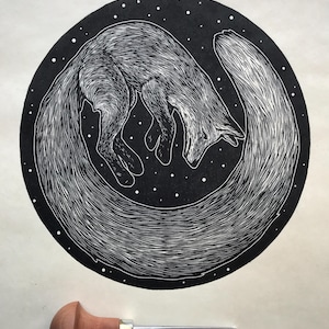 May include: A black and white linocut print of a fox curled in a circle, set against a starry background. The fox's fur is detailed with fine lines. A wood-handled linocut tool rests below the print.