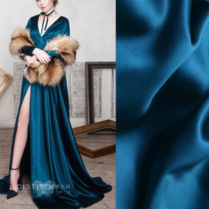 Puede incluir: Un vestido de terciopelo azul teal con una falda larga y fluida y una abertura alta. El vestido tiene un escote en V y está adornado con una estola de piel marrón. La tela es suave y lujosa.