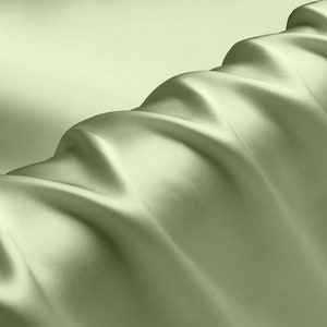 Puede incluir: Una tela suave de color verde claro con una textura suave y fluida. La tela está drapeada en pliegues, creando un aspecto lujoso y elegante.